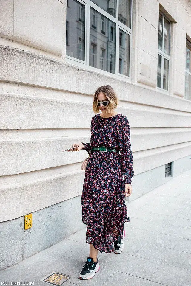 Petite woman in a dark navy long-sleeve midi dress with small scattered pink and red florals, cinched at the waist with a dark green embellished belt, wearing chunky dark sneakers with pink accent details, carrying a small blush crossbody bag, and white cat-eye sunglasses, standing on a city pavement beside a stone building.