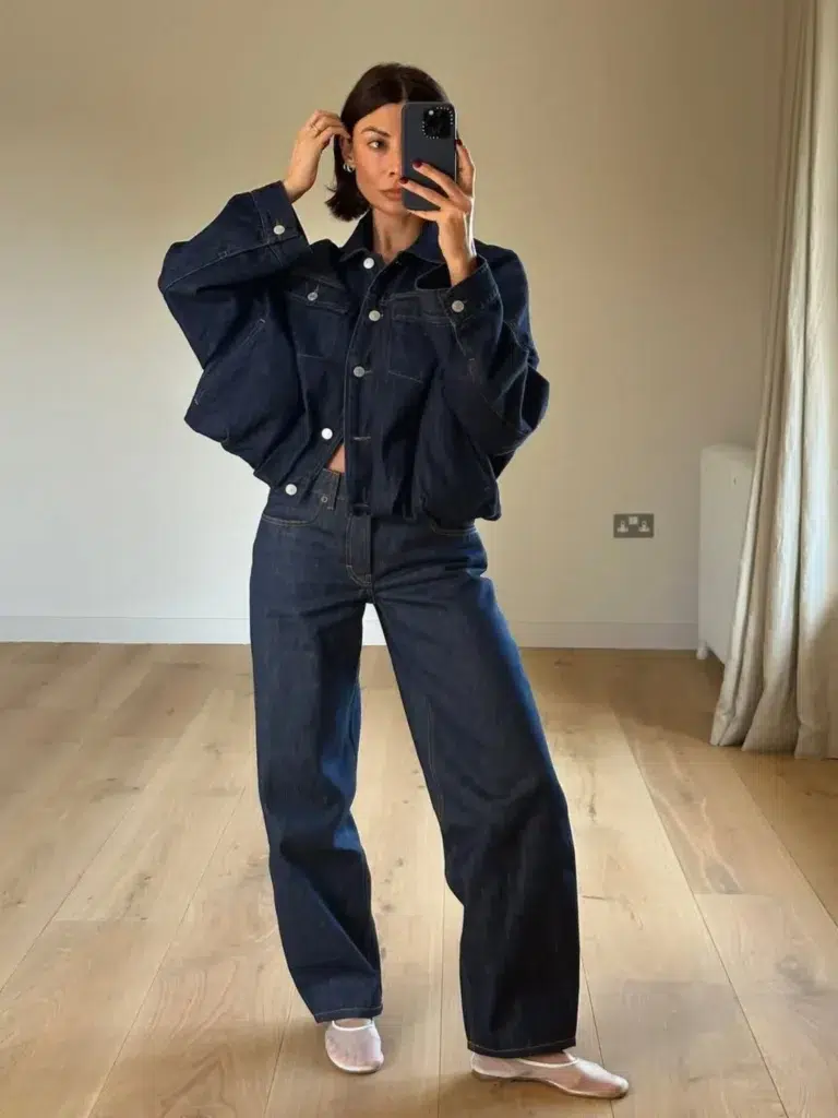 Petite woman in a matching dark indigo denim co-ord set consisting of an oversized cropped denim jacket with silver buttons and structured volume at the shoulders, paired with wide-leg dark indigo jeans in the same wash, wearing white slipper-style ballet flats, taking a mirror selfie in a minimalist room with warm wood floors and a neutral-toned wall.