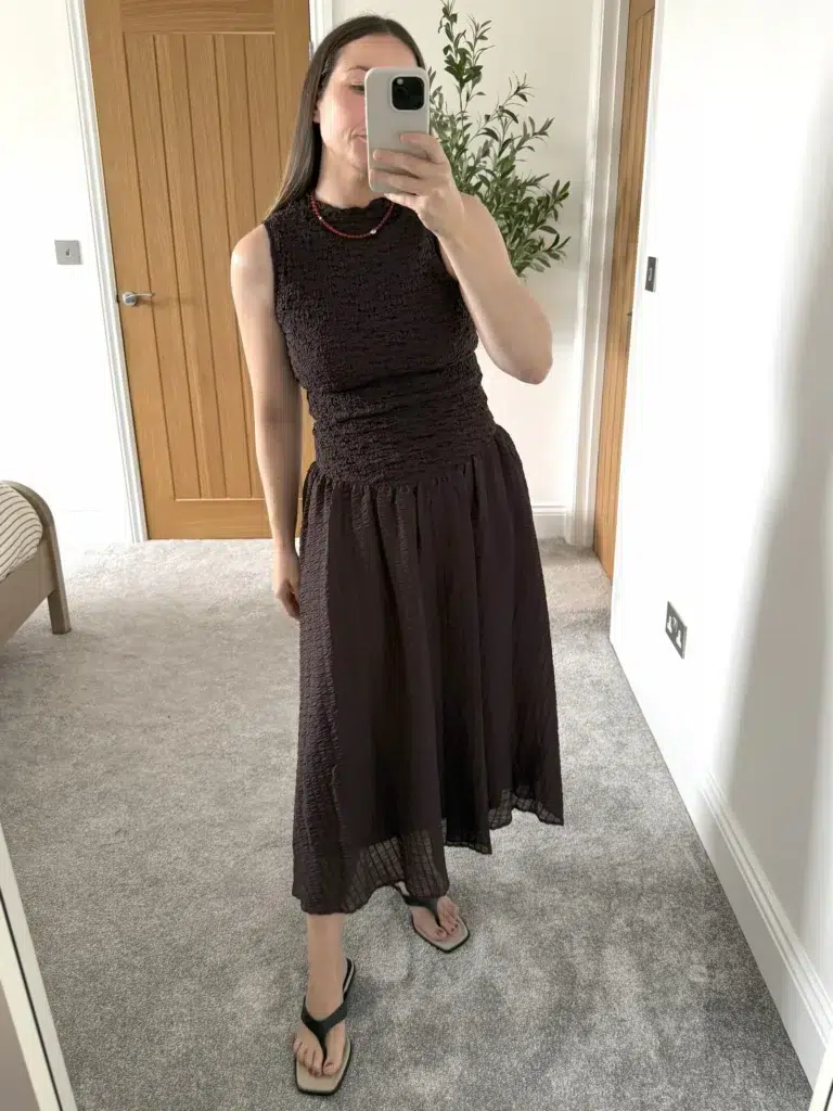 Woman taking a mirror selfie in a deep chocolate brown sleeveless smocked midi dress with a defined elasticated waist, soft crinkled flowy skirt hitting at mid-calf, paired with dark flat thong sandals and a small red beaded necklace, photographed in a carpeted bedroom with a wooden door in the background.