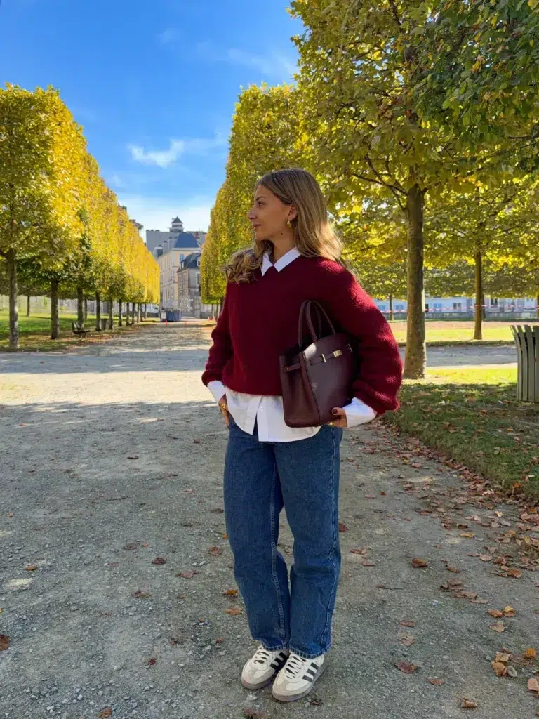 Petite woman in a deep crimson oversized chunky knit sweater layered over a white button-down Oxford shirt with the white collar and shirt hem both visible, paired with medium-wash relaxed straight blue jeans and retro white and black sneakers, carrying a structured oxblood leather tote bag with gold hardware tucked under her arm, standing on a gravel path in a French-style park lined with golden autumn trees.