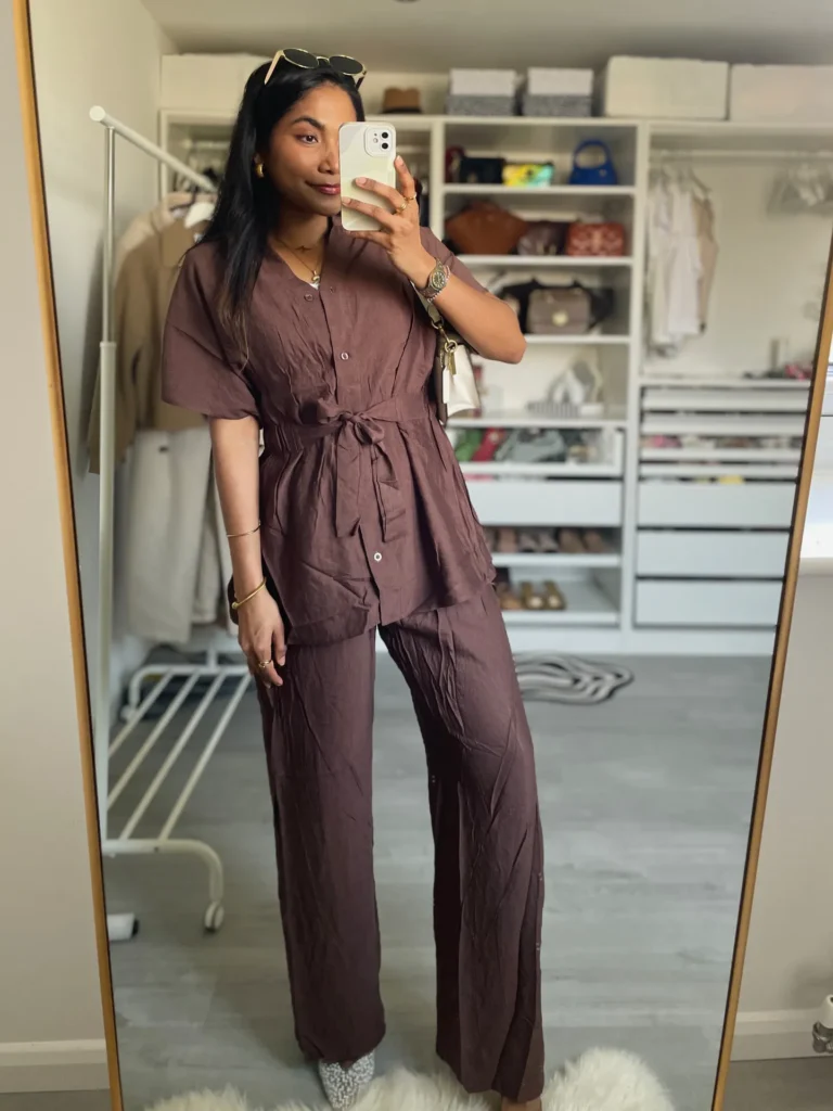 Petite woman in a deep plum crinkled linen matching co-ord set with a short-sleeve button-down top tied at the waist with a self-sash belt and wide-leg full-length pants, taking a mirror selfie in a walk-in closet with organized white shelving visible in the background.