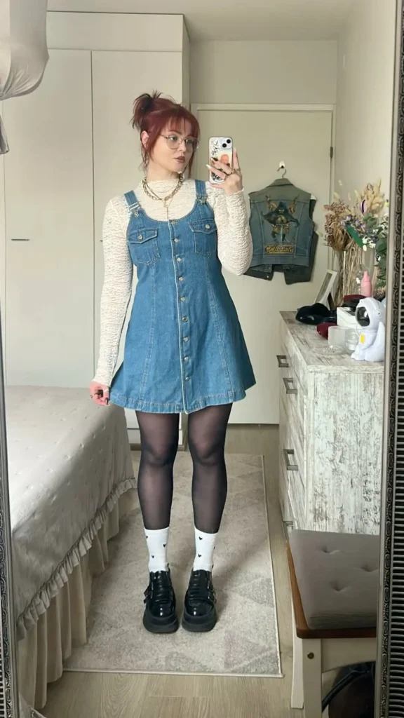 Woman taking a mirror selfie in a medium blue wash wide-strap button-front denim pinafore mini dress with chest flap pockets and a flared skirt, worn over a cream textured long-sleeve fitted top with full sleeves visible at the wrist, styled with black semi-sheer tights, white heart-print crew socks folded at the calf over the tights, chunky black platform buckle Mary Jane shoes, layered gold chain necklaces, and round thin wire-frame glasses, with auburn-red hair in a messy top bun with bangs, photographed in a bedroom with a vintage white dresser and a denim vest hanging in the background.