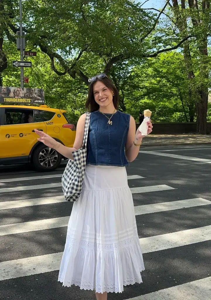 Petite woman standing on a crosswalk in a city park, wearing a fitted sleeveless denim vest tucked at the waist over a long white tiered eyelet prairie skirt, carrying a navy gingham tote, holding an ice cream cone, with gold jewelry and a yellow taxi behind her.