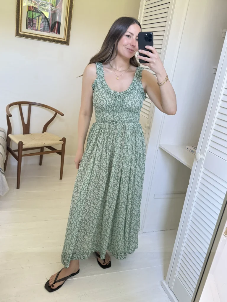 Woman taking a mirror selfie in a bright white interior room in a sage green and cream small ditsy floral dress with ruffled shoulder straps, a scoop neckline with a small center tie detail, a smocked elasticated waist section, and a full gathered skirt hitting at the lower calf, paired with black square-toe flat thong sandals, a delicate gold coin pendant necklace, and a thin gold chain bracelet.