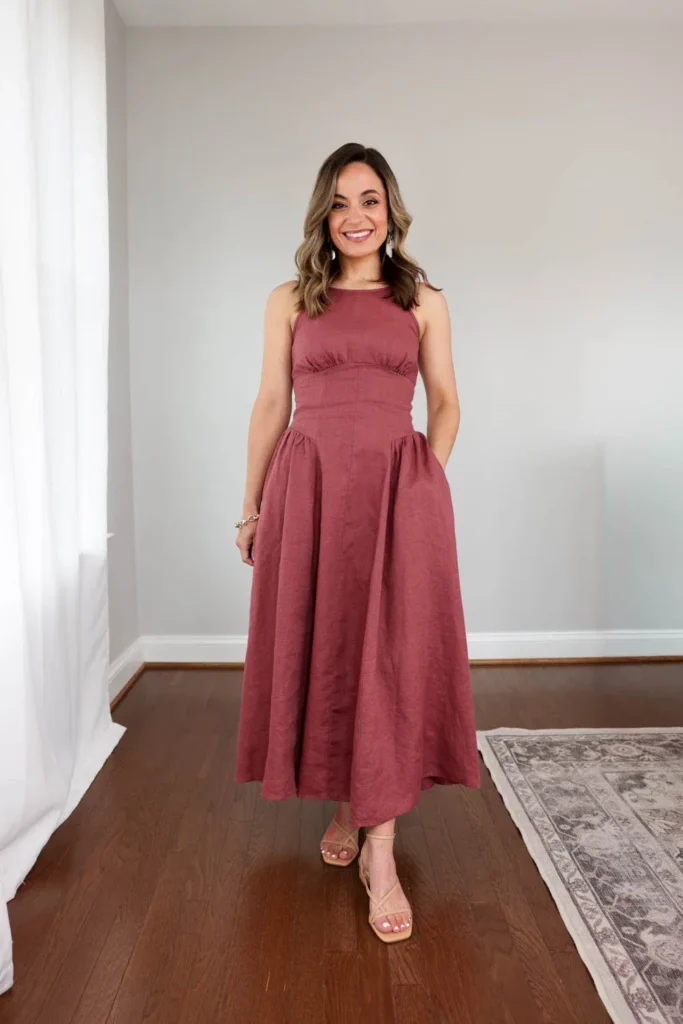 Smiling woman standing in a bright indoor room in a dusty rose mauve sleeveless linen empire waist midi dress with a structured underbust band, high round neckline, and full gathered A-line skirt hitting at mid-calf, one hand in the dress pocket, paired with gold strappy lace-up square-toe heeled sandals, cream teardrop drop earrings, and a slim gold chain bracelet, with hardwood floors and a grey patterned area rug visible.