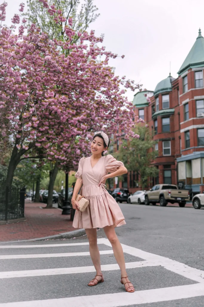 Petite Asian woman in a dusty rose blush wrap mini dress with short puff sleeves, a V-neckline, side self-tie waist, and a voluminous tiered gathered skirt falling above the knee, wearing flat tan multi-strap sandals, carrying a tiny structured woven rattan mini clutch, and wearing a pink floral headband, standing on a pedestrian crosswalk in an urban neighborhood with a cherry blossom tree in full pink bloom and red brick apartment buildings in the background.
