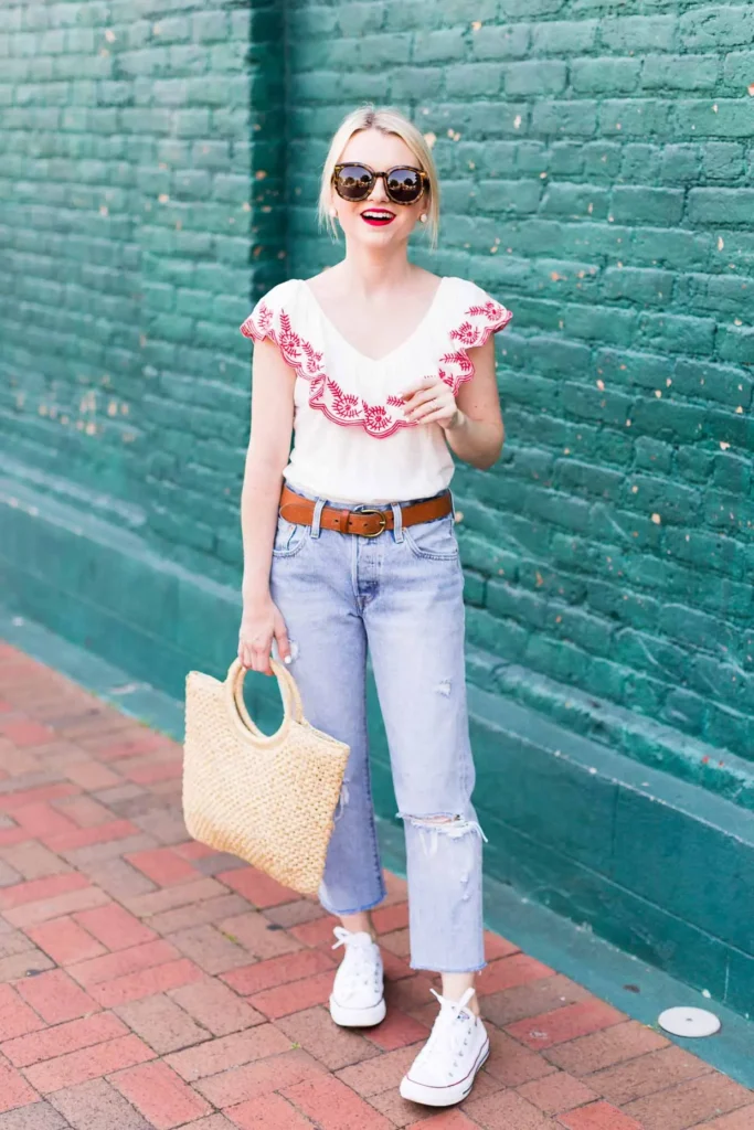 Woman in light wash distressed high-waisted mom jeans with a raw frayed hem, a white flutter-sleeve top with red scalloped embroidery around the neckline, a cognac leather belt, white high-top Converse sneakers, and a natural woven straw tote with circular ring handles, standing against a teal painted brick wall, wearing red lipstick and tortoiseshell sunglasses