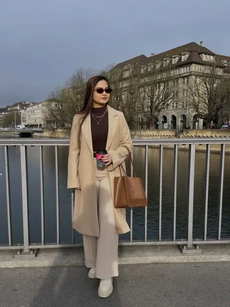 Petite woman with long straight dark hair standing on a riverside bridge in a European city with a grey winter sky and stone buildings across the water, wearing an open longline camel wool coat over a fitted deep espresso brown ribbed turtleneck, cream wide-leg tailored trousers running to the floor, cream chunky lug-sole ankle boots in the same tone as the trousers, layered delicate gold necklaces at the collar, small oval dark sunglasses, and carrying a structured cognac tan leather tote bag while holding a branded coffee cup.