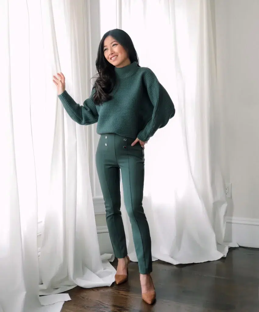 Petite Asian woman with long dark hair smiling in a bright white room with sheer floor-length white curtains and dark hardwood floors, wearing a forest green chunky ribbed turtleneck sweater with balloon sleeves cropped at the high waist, matching forest green high-rise slim-fit tailored trousers with a front pintuck seam and silver button detail at the waistband, and tan camel pointed-toe stiletto pumps.