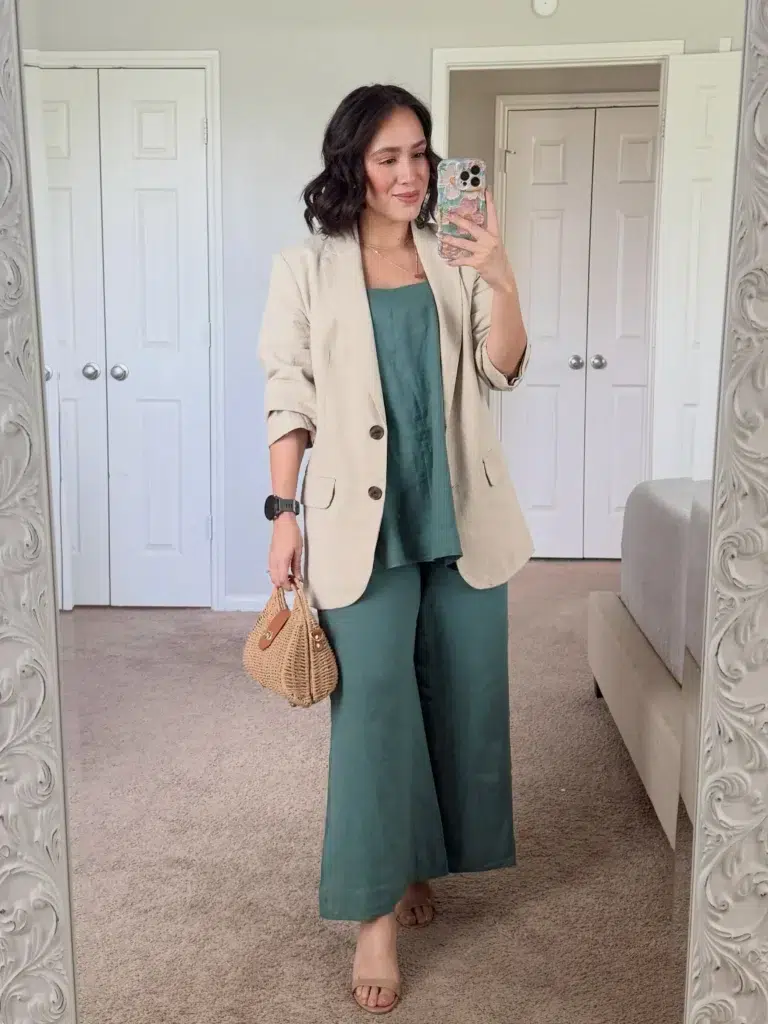 Curvy petite woman taking a bedroom mirror selfie wearing a forest green linen wide-leg pants and square-neck camisole co-ord set layered under an oversized natural oatmeal linen blazer with rolled sleeves and button detail, styled with flat tan slide sandals, a small structured woven rattan basket bag, a black watch, and delicate layered gold necklaces.