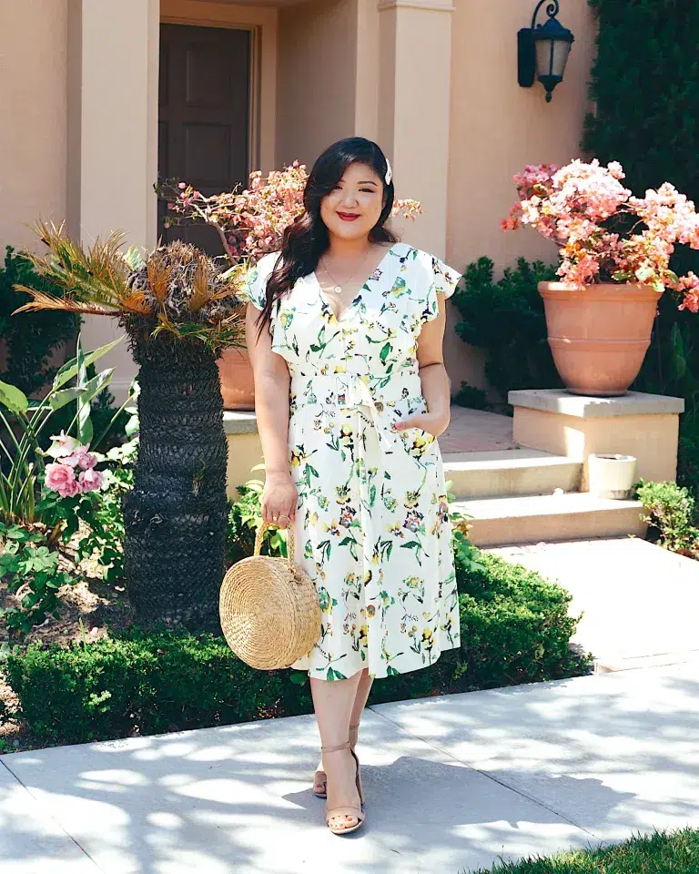 Curvy petite woman standing outdoors in an ivory floral cap sleeve midi dress with a cinched waist tie, wearing nude strappy block-heel sandals and carrying a round woven straw bag with bamboo handles, styled with a white floral hair clip, delicate gold necklace, and bold red lip, in front of a Mediterranean-style home with flowering bougainvillea in terracotta pots.