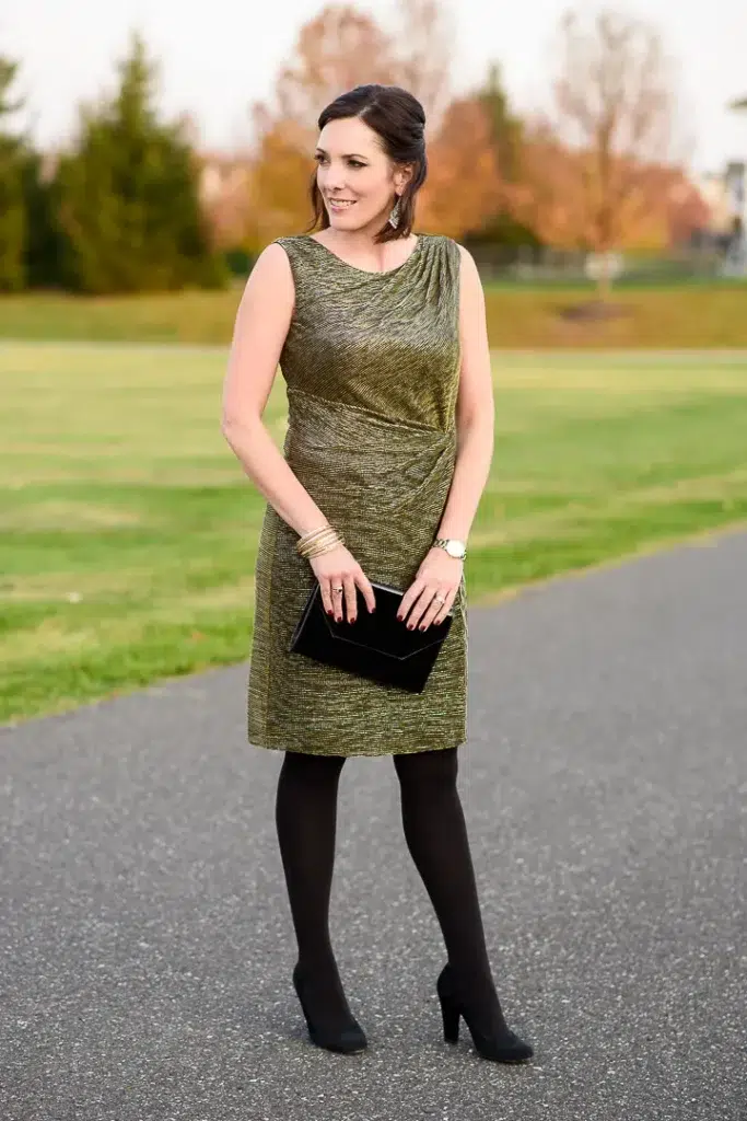 Petite woman in a gold and olive metallic sleeveless sheath dress with a draped cowl neckline and side ruching, styled with black opaque tights, black matte block-heeled pumps, a black geometric envelope clutch, and stacked gold bangles, standing on an outdoor path with autumn trees behind her.