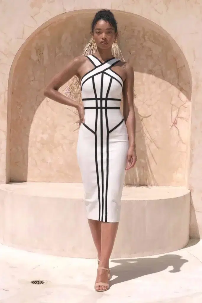 Slender woman with dark curly hair pulled into a high bun wearing a white bandage bodycon midi dress with bold black graphic vertical stripe detailing and a criss-cross halter neckline, paired with nude strappy heeled sandals and large gold fringe tassel drop earrings, standing in front of a curved sandy terracotta archway wall in bright outdoor daylight.