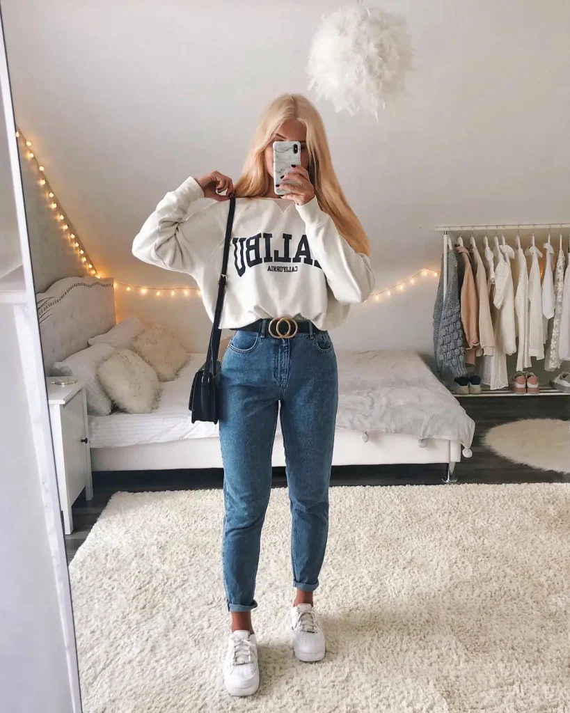 
Young woman taking a bedroom mirror selfie in
medium wash high-waisted mom jeans with a single ankle cuff, a white graphic sweatshirt, a double-ring gold buckle tan belt worn over the sweatshirt, white
chunky platform sneakers, and a small black
crossbody bag, in a cozy white bedroom with fairy lights