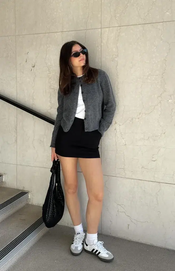 Petite woman leaning against a marble wall by a staircase, wearing a charcoal grey cardigan over a white tee, a short black mini skirt, white crew socks, and grey-and-white low sneakers, holding a soft black woven bag, with narrow black sunglasses.