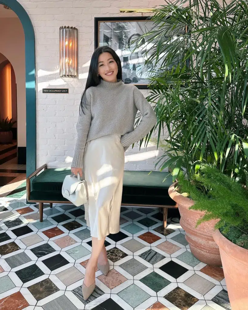 Petite woman standing in a decorative hotel lobby with geometric marble tile floors and a green velvet bench, wearing a cropped grey chunky textured knit mock-neck sweater landing at the waistband of a champagne cream bias-cut satin midi skirt, with a brief flash of dark ankle socks visible above pointed nude mule heels, holding a small structured white top-handle bag, long dark hair worn down, small gold hoop earrings.
