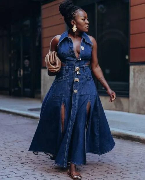 Woman in a dark navy indigo-wash sleeveless denim halter maxi dress with a shirt collar, deep plunging V-neckline, decorative gold hardware buttons and buckles down the center front placket, side waist cut-outs, and a full flared skirt with dual deep front slits, styled with large sculptural gold flower earrings, a tan soft slouchy pouch shoulder bag, flat tan strappy sandals, and hair piled into a high bun, walking on a brick-paved city street with a terracotta building backdrop.