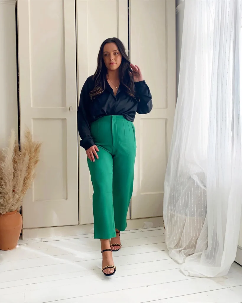 Woman wearing a loose black satin button-down shirt tucked loosely into high-waisted bright green wide-leg cropped trousers with front seam detail, paired with black strappy heeled sandals with gold chain hardware, indoors against a white backdrop — a bold color petite interview outfit idea for creative industries