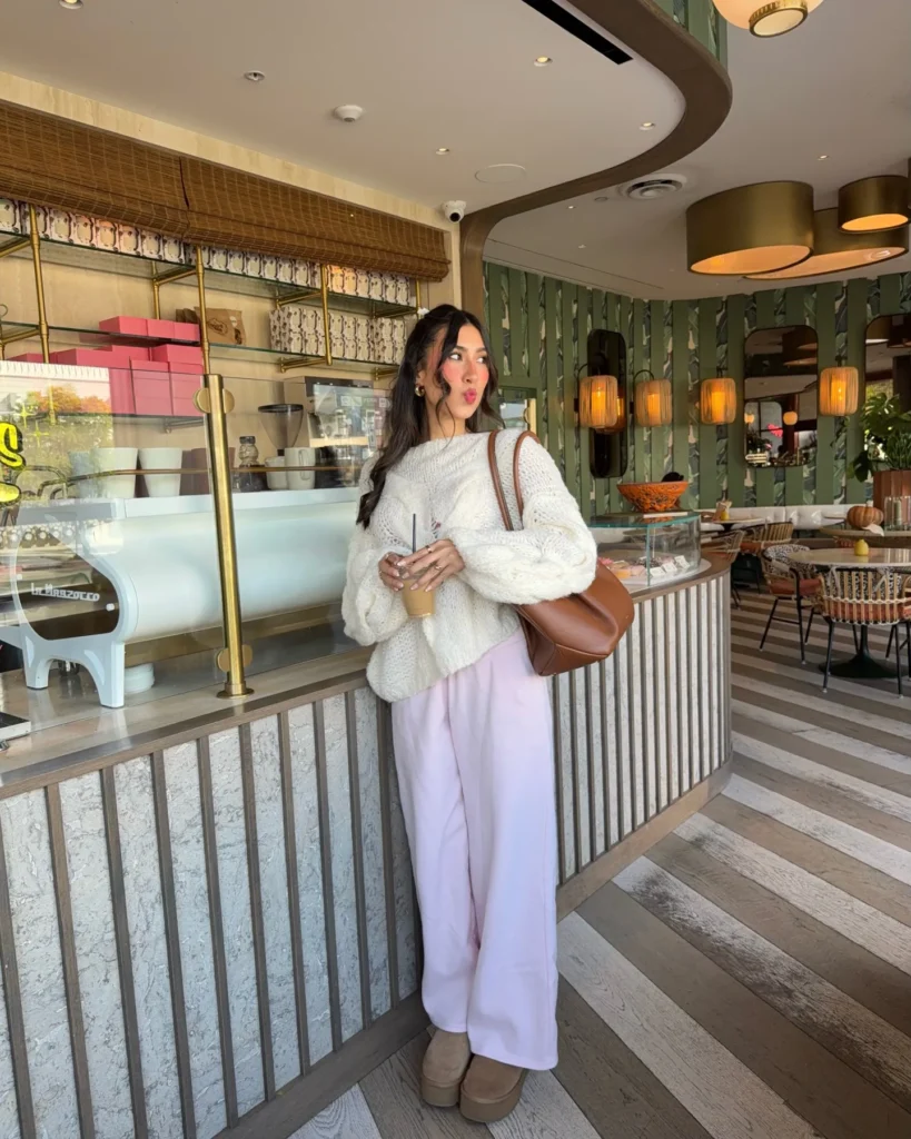 Petite woman with long dark wavy hair leaning against a ribbed counter inside a stylish café with green botanical wallpaper and warm pendant lighting, wearing a chunky open-weave cream ivory oversized knit sweater landing at the hip, pale lavender wide-leg soft trousers running to the floor, tan camel platform UGG-style mini boots, gold hoop earrings, and a rounded cognac brown shoulder bag, holding an iced coffee drink.