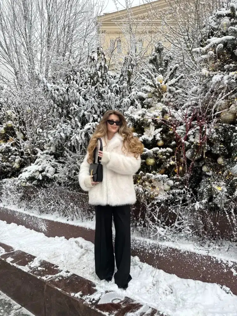 Petite woman with long voluminous curly golden-blonde hair standing outdoors in a snowy winter scene with snow-covered Christmas trees decorated with gold ornaments behind her, wearing a cropped ivory white fluffy faux fur jacket with a wide lapel collar and cream faux fur cuffs at the wrists, black wide-leg tailored trousers with a center crease running full length to the ground, small black cat-eye sunglasses, and holding a compact black structured top-handle bag with gold hardware against her chest.
