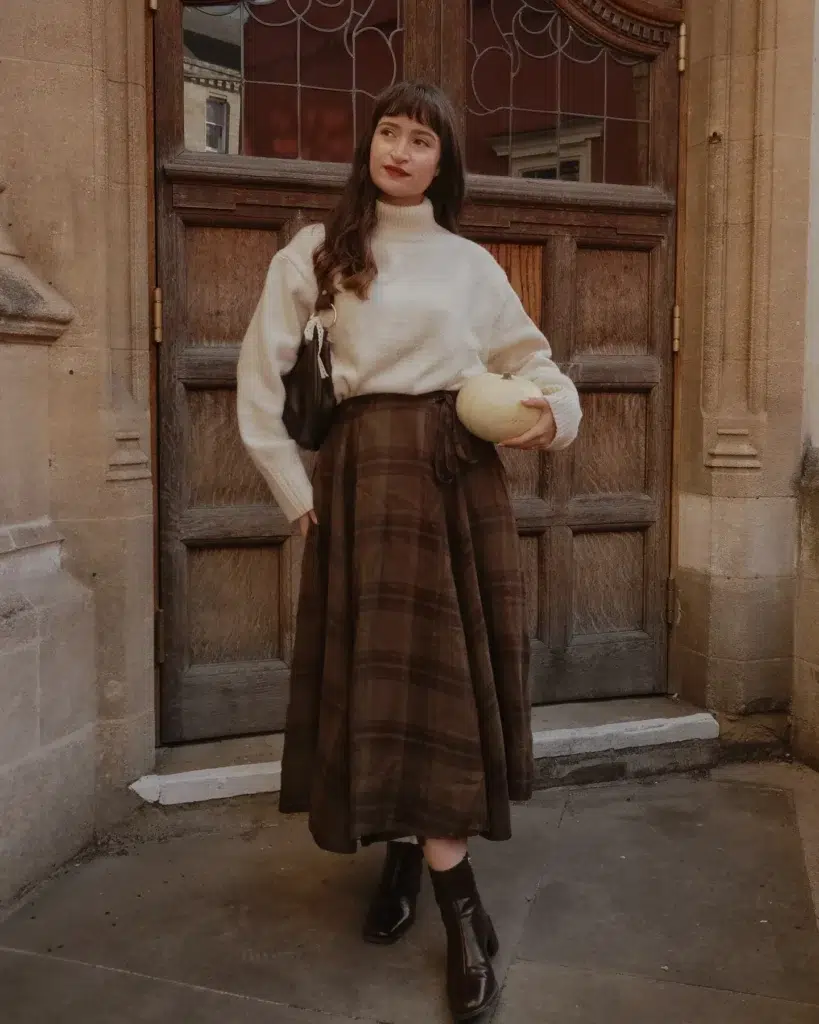 Petite woman in a cream ivory oversized turtleneck sweater with wide voluminous sleeves, cleanly tucked into a deep chocolate brown tonal plaid wrap midi skirt with a visible self-tie knot at the hip, paired with dark chocolate brown square-toe ankle boots, carrying a small dark brown crescent-shaped shoulder bag with a gold ring strap detail, wearing a red lip, standing in front of an ornate dark carved wooden door with decorative glass pane details and stone columns of a historic British stone building, holding a small pale cream gourd.