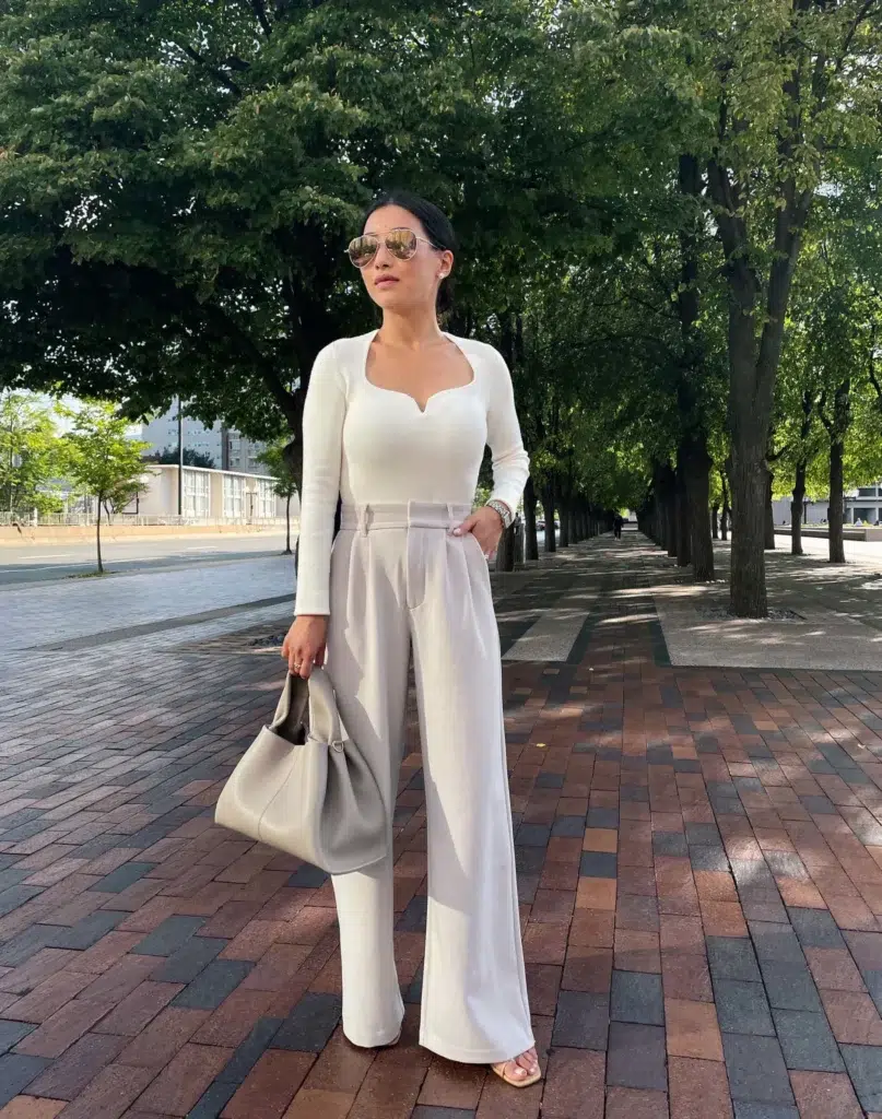 Petite woman in high-waist light stone wide-leg trousers and a fitted cream long-sleeve top with a notched neckline, carrying a soft sage leather hobo shoulder bag, wearing nude strappy heeled sandals while walking along a tree-lined European promenade.