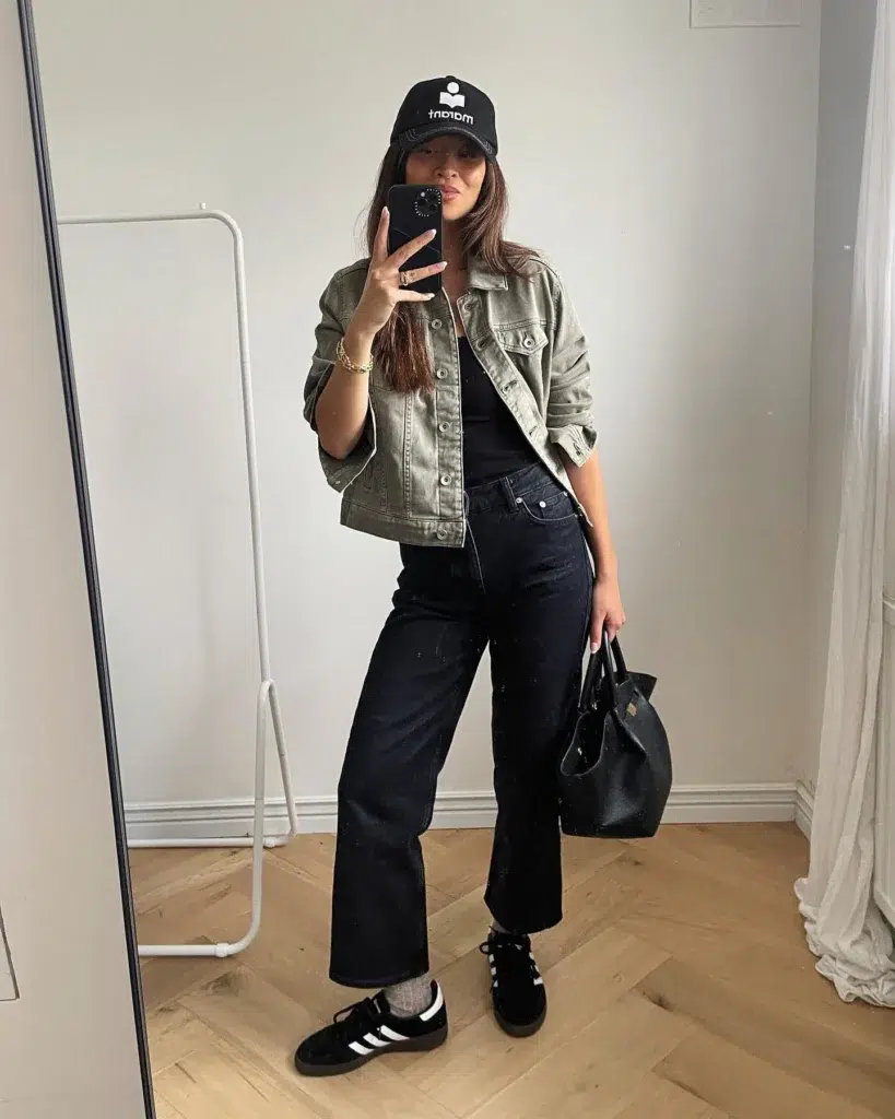 Petite woman taking a mirror selfie in a minimal white room wearing high-waisted black straight leg jeans, a fitted black top, a washed olive green cropped denim jacket with snap buttons and chest pockets, black suede low-profile sneakers with white side stripes and a gum rubber sole, grey ankle socks, a black baseball cap with a small white logo, and a structured black leather tote bag with gold hardware carried at her side, with a gold chain bracelet on her wrist.