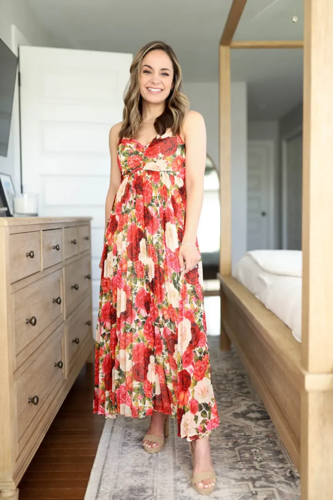 Petite woman with shoulder-length wavy brunette hair in a cream-base large red and pink oversized rose floral empire-waist maxi dress with thin straps, ruched bust detail, and a full tiered skirt, wearing nude open-toe platform wedge sandals and a delicate bracelet and fine chain necklace, smiling indoors in a bright bedroom with a wood dresser and wood canopy bed frame visible in the background.