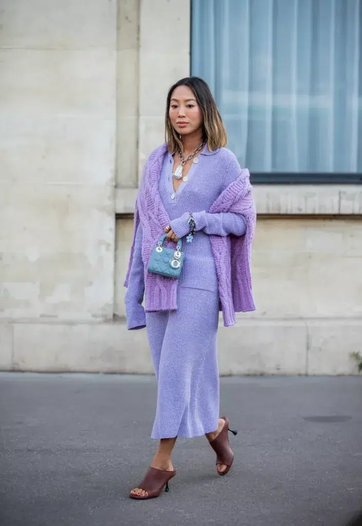 Petite Asian woman with a chin-length bob wearing a soft lavender fine ribbed polo collar midi sweater dress with a slim fitted silhouette, layered with a matching lavender oversized chunky cable knit cardigan draped off both shoulders, paired with deep burgundy leather open-toe mule heels, a tiny teal quilted micro handbag, and layered silver chain and coin necklaces, standing on a street pavement in front of a pale stone building facade with large glass windows.