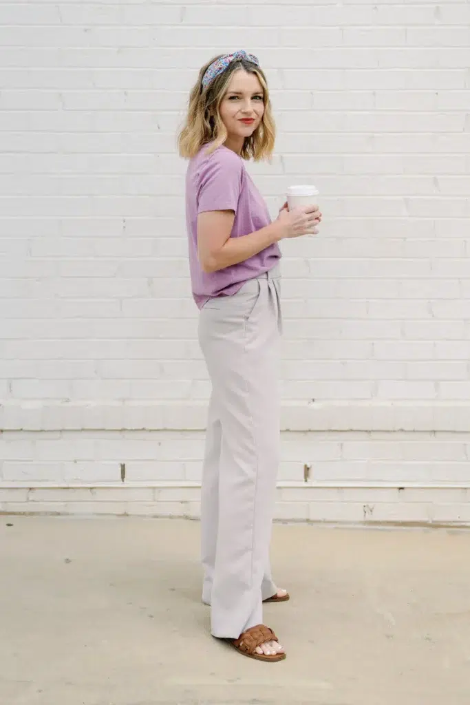 Petite woman shot from the side in a short-sleeve soft lavender t-shirt tucked into high-rise light grey-lavender wide-leg trousers with a self-tie paper bag bow waist, wearing cognac woven flat slide sandals and a floral knotted headband, holding a white takeaway coffee cup, standing against a white painted brick wall.