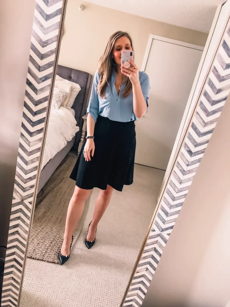Petite woman in a mirror selfie wearing a light blue v-neck blouse with rolled sleeves and a black A-line knee-length skirt, with black studded pointed-toe heels and a delicate watch — a business casual petite interview outfit idea