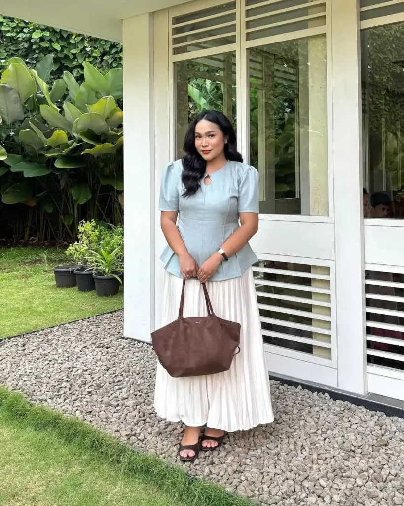 Petite pear-shaped woman in a fitted light blue structured peplum top with short sleeves and a keyhole neckline detail, paired with a cream accordion-pleated maxi skirt, wearing dark brown flat sandals and carrying a brown leather tote bag, standing outside a modern white building surrounded by lush tropical greenery.