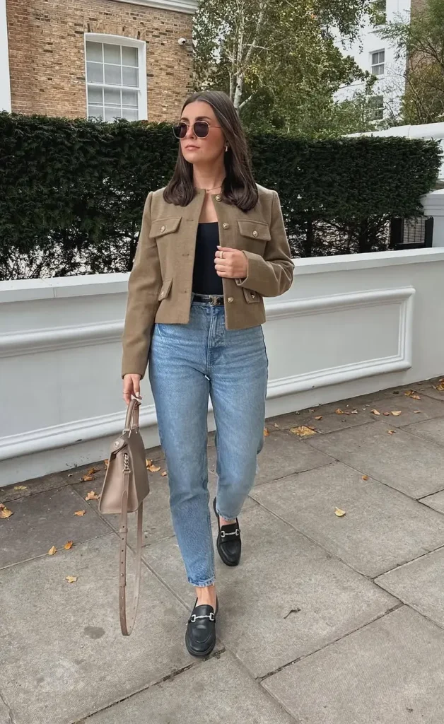 Petite woman wearing light wash ankle-length mom jeans, a cropped olive military jacket with gold buttons, a black fitted top, and black horsebit loafers, carrying a nude structured bag on a London street