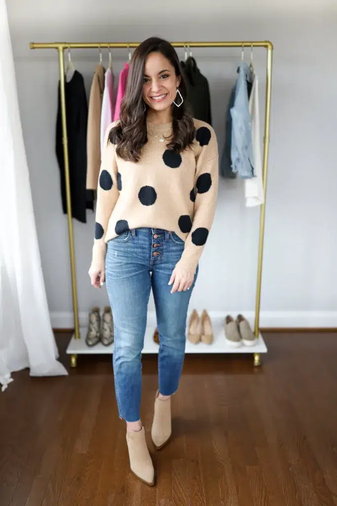 Petite woman standing in front of a gold clothing rack wearing high-waisted light wash straight leg jeans with an exposed copper button fly and a raw frayed ankle hem, a camel crew-neck knit sweater with large oversized navy polka dots grazing the waist, tan suede pointed-toe ankle boots, large geometric white statement earrings, and layered gold chain necklaces.