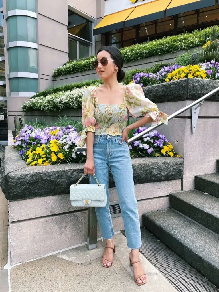 Petite woman standing on outdoor stone steps wearing high-waisted light wash straight leg jeans with a raw frayed cropped hem, a floral smocked crop top with a square neckline and dramatic voluminous puff sleeves in a yellow pink and green floral print, minimal tan strappy block-heel sandals, and a small pale mint quilted flap bag with a gold chain strap and gold hardware, with a low chignon updo and gold-frame aviator sunglasses.
