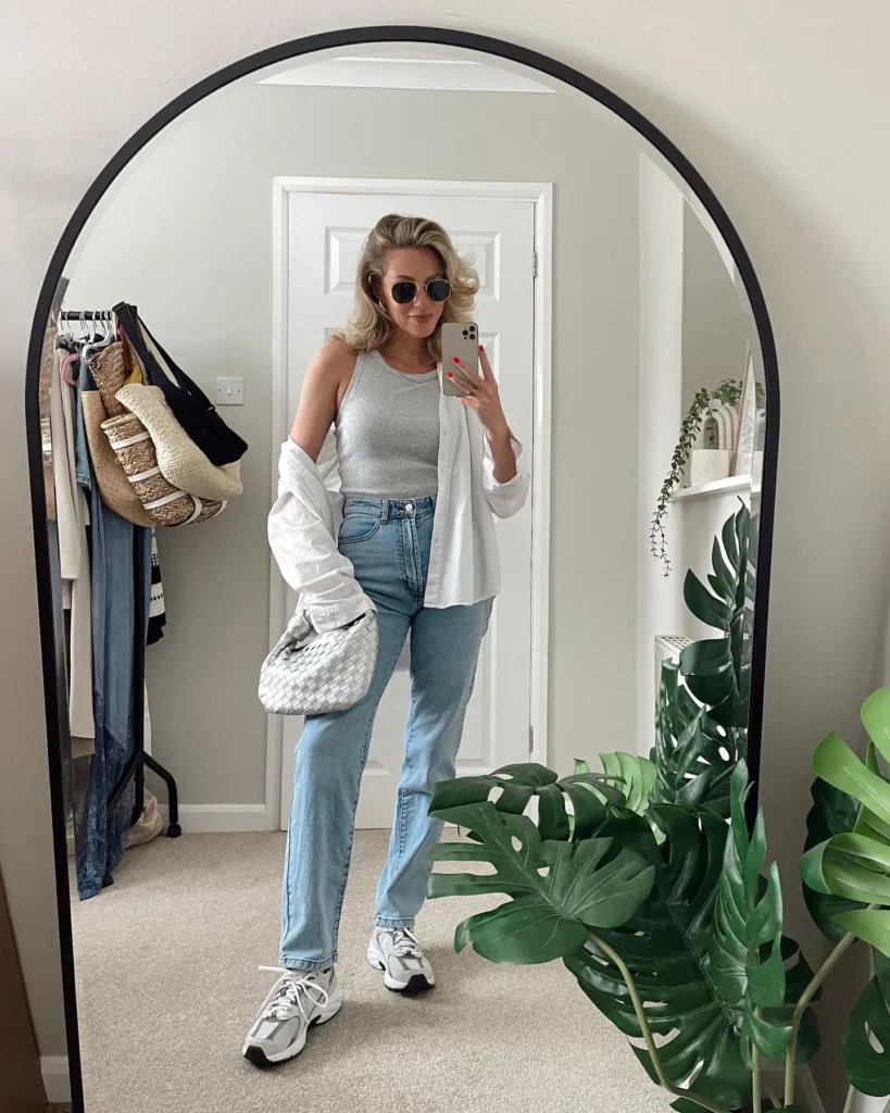 Petite woman taking a mirror selfie in a bedroom wearing high-waisted light wash pale blue straight leg jeans, a grey ribbed racerback tank top, a white oversized button-down shirt worn open as a layering piece, white chunky dad-style sneakers, and a small white woven textured hobo bag, with small oval sunglasses and voluminous wavy blonde hair.