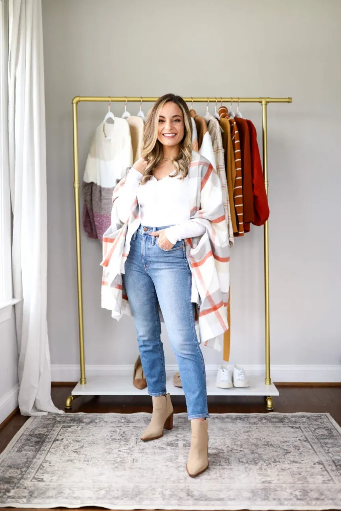 Petite woman standing in a bright home interior in front of a gold clothing rack wearing high-waisted light wash straight leg ankle-length jeans, a fitted white long-sleeve top, an oversized cream plaid blanket scarf with rust and grey grid lines draped open over her shoulders, and tan suede block-heel ankle boots, with layered delicate gold necklaces at the neck.