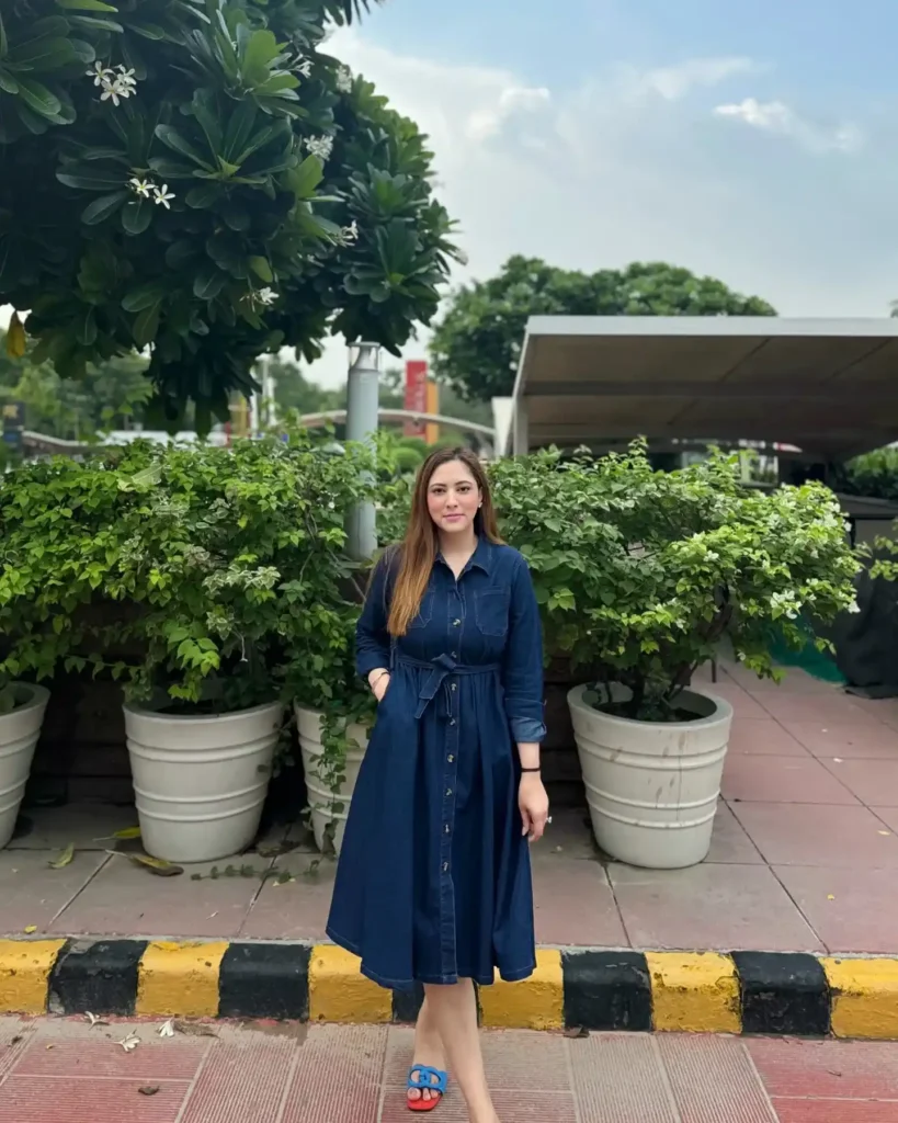Woman in a dark navy wash long-sleeve button-front denim shirt dress with a pointed collar, gold buttons down the center placket, chest patch pockets, a self-tie fabric belt creating a gathered waist, and a full A-line midi skirt hitting below the knee, styled with bright cobalt blue flat slide sandals and a black fitness tracker on the wrist, standing outdoors on a paved walkway in front of large white planters with lush green trees and foliage.