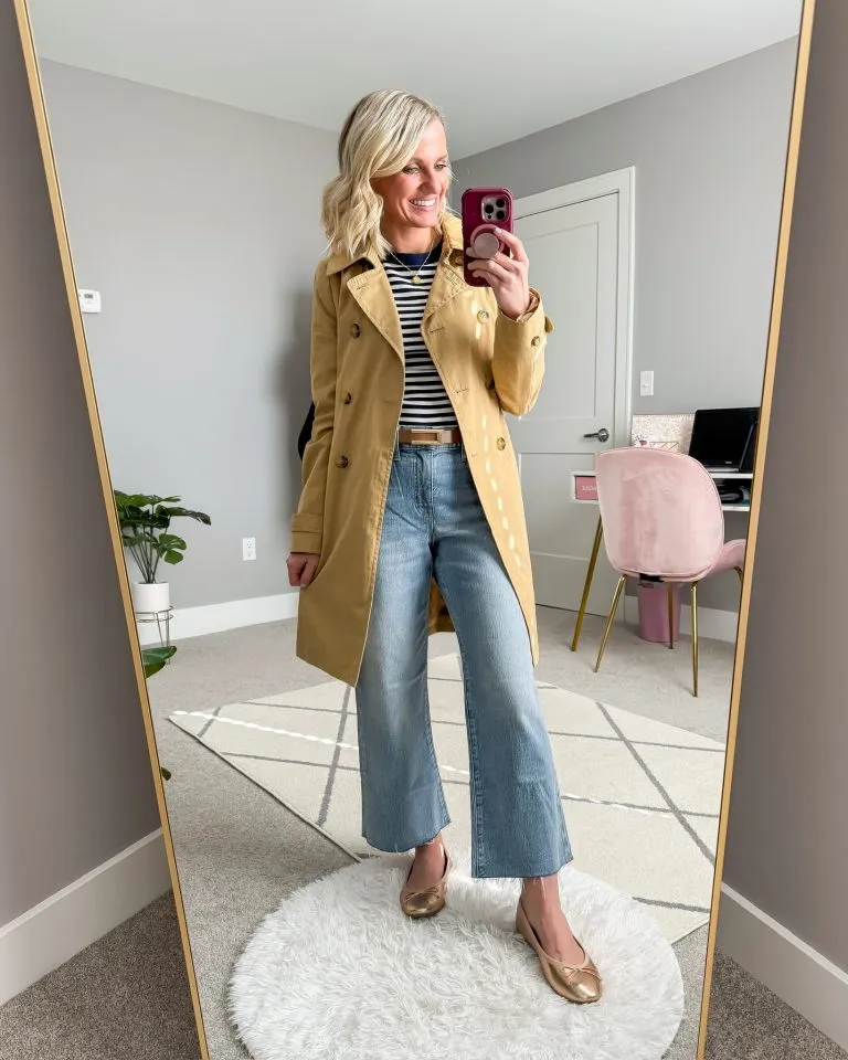 Petite woman wearing medium-wash wide-leg jeans with a striped top, tan trench coat belted at the waist, and gold ballet flats showing how to wear a longer layer over wide-leg jeans by defining the waist with a belt