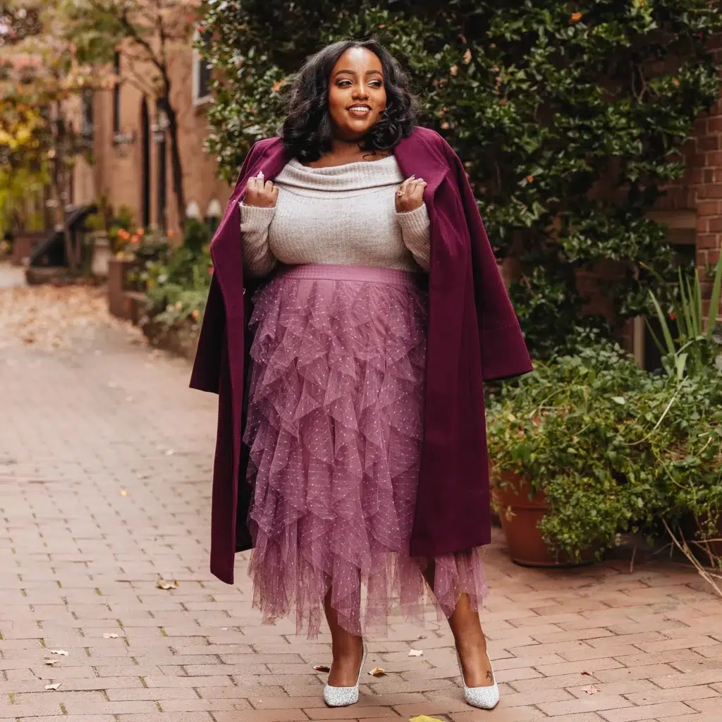 Petite plus-size woman with short dark curly hair wearing a deep plum wine longline wool coat worn open and draped at the lapels over a soft gray off-shoulder cowl-neck ribbed knit sweater and a mauve dusty rose cascading tiered asymmetric tulle ruffle midi skirt with a subtle polka dot texture throughout, paired with silver glitter pointed-toe pumps, a thin dark choker necklace and rings, walking on a brick-paved autumn street lined with brick buildings, green hedges, terracotta planters, and scattered fallen leaves.