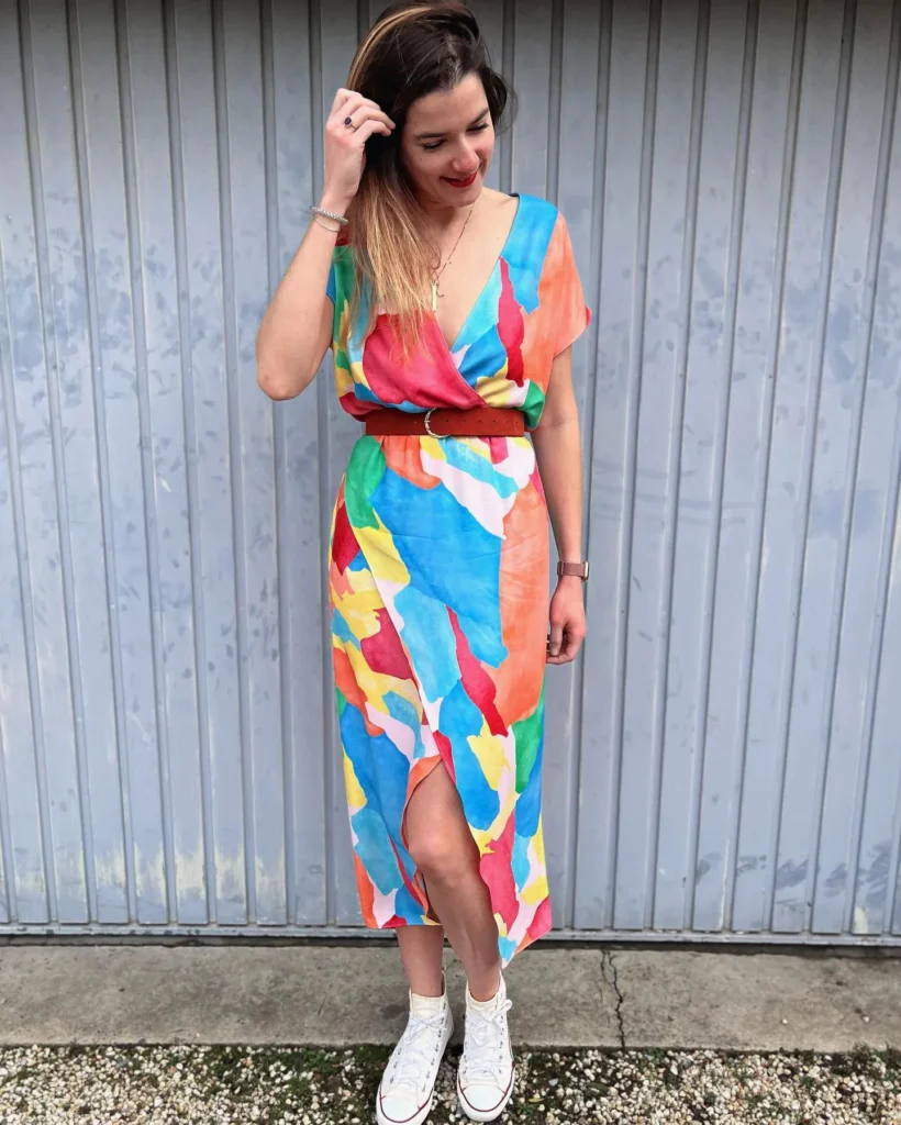 Woman in a deep V-neck wrap midi dress in a bold abstract brushstroke print with large color-block splashes of cobalt blue, coral orange, hot pink, yellow, and green on a white ground, with short kimono-style drop shoulder sleeves and a front wrap slit at the hem, styled with a separate cognac rust leather belt with a silver ring buckle layered at the natural waist, white high-top canvas sneakers, a delicate gold chain necklace, a silver bracelet, and a deep red lip, standing against a pale corrugated blue-grey metal wall.