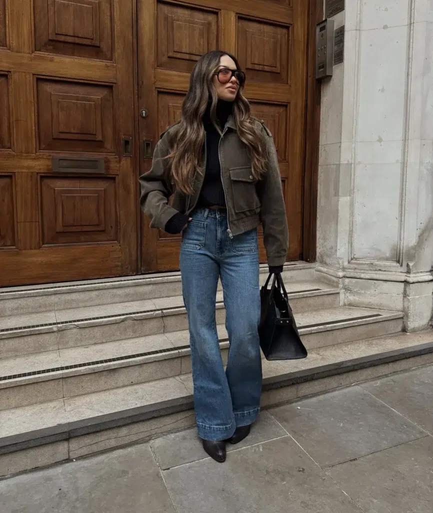 Petite woman in medium wash wide-leg flare jeans and a cropped olive khaki bomber jacket with a ribbed waistband, layered over a fitted black turtleneck, styled with black pointed-toe ankle boots, a large structured black leather tote, and oversized round amber-tinted sunglasses on stone steps outside a grand European-style building.