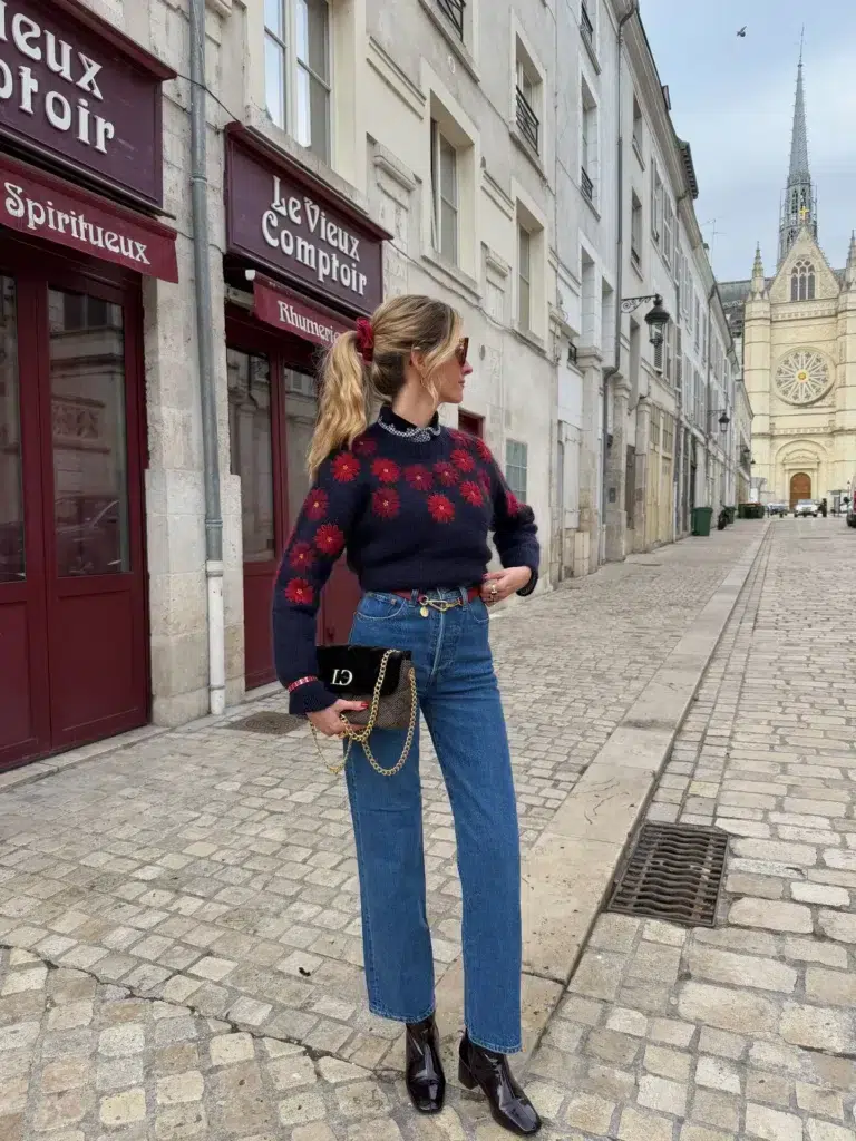 Petite woman standing on a cobblestone French street wearing high-waisted medium-wash blue straight leg jeans, a chunky navy knit sweater with an all-over red floral daisy pattern, a black collared shirt with a sparkly embellished collar layered underneath, oxblood patent leather block-heel square-toe ankle boots, a small black velvet mini bag with a gold chain strap, a thin waist belt with a small gold charm pendant, tortoiseshell sunglasses, and a red scrunchie holding a low ponytail.