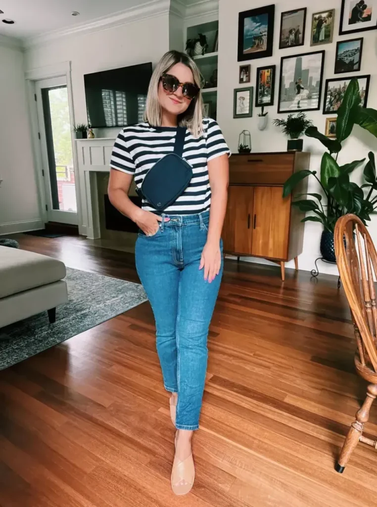 Petite woman standing in a bright living room with a gallery wall behind her, wearing high-waisted medium wash blue straight leg cropped jeans, a short-sleeve bold navy and white horizontal stripe crew-neck t-shirt, a navy blue crossbody belt bag worn diagonally across the chest, and nude flat open-toe mule slides, with large tortoiseshell sunglasses and a blunt blonde bob.