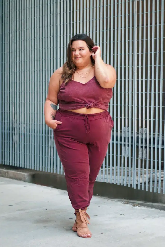 Petite plus-size woman in a mineral-washed deep burgundy two-piece matching set, wearing a lace-trim V-neck front-tie crop cami and tapered drawstring jogger pants with side pockets, styled with tan lace-up flat wrap sandals and layered delicate gold necklaces, standing on a city sidewalk in front of a vertical metal slat wall.