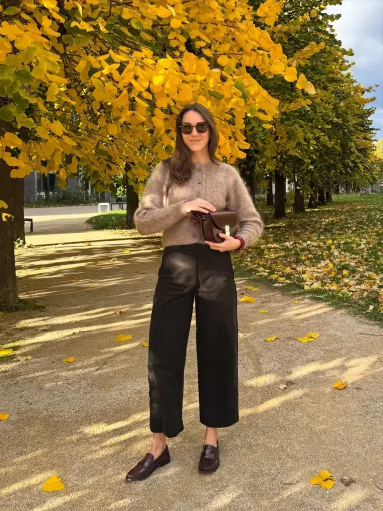 Petite woman in a fluffy mohair-style cropped cardigan in a warm mink-mushroom tone with a button-front and crew neck, paired with high-waisted wide-leg cropped black trousers hemmed to show the full shoe, wearing dark oxblood burgundy textured penny loafers, holding a small structured dark oxblood leather satchel bag with a gold clasp, wearing dark tortoiseshell sunglasses and a beaded bracelet on her wrist, standing on a gravel path in a formal park lined with golden yellow autumn trees with scattered fallen leaves on the ground.