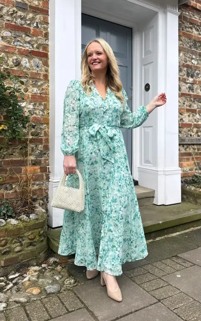 Woman wearing a mint and teal watercolor floral chiffon wrap maxi dress with long blouson sleeves, a V-neckline, and a self-tie waist bow, paired with nude block-heeled pumps and a white pearl top-handle mini bag outside a cottage setting.