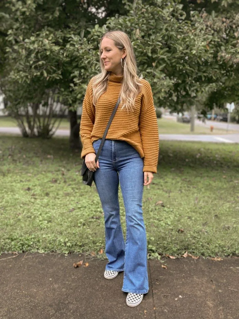 Petite woman in medium wash flare jeans and a mustard yellow ribbed mock-neck sweater, styled with checkerboard Vans and a dark mini crossbody bag for a retro 70s look.