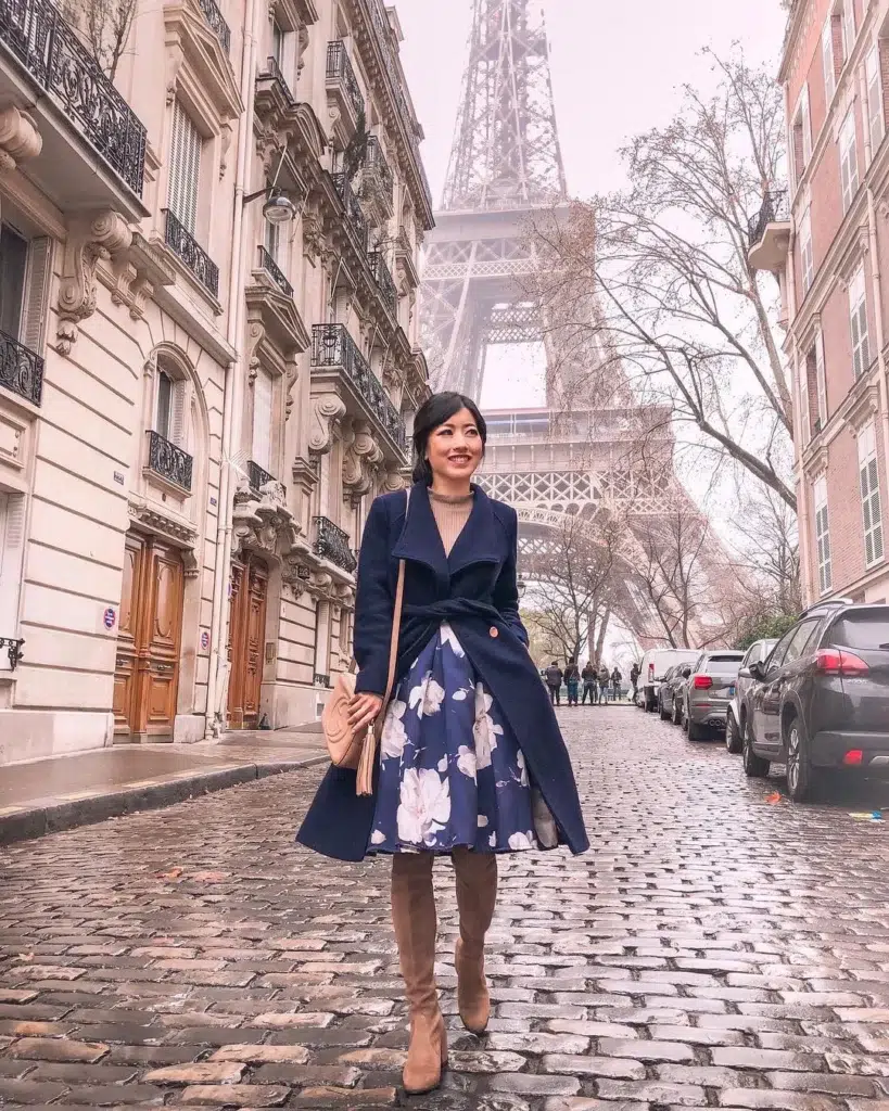 Petite woman in a navy belted wrap coat tied at the natural waist, worn over a navy and white large floral A-line midi skirt, with a slim camel turtleneck visible at the neckline, tall camel suede knee-high boots, and a blush tassel crossbody bag, standing on Parisian cobblestones with the Eiffel Tower in the misty background.