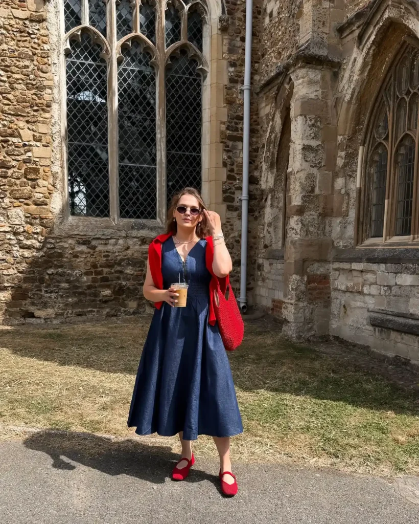Woman in a dark navy sleeveless denim V-neck midi dress with a front zip detail and full flared A-line skirt, styled with a red cardigan draped over both shoulders, a red open-weave woven tote bag, red suede Mary Jane flat shoes, small oval tinted sunglasses, a delicate silver chain necklace, a silver bracelet, and small drop earrings, holding an iced coffee and standing on a sunlit stone path beside a historic Gothic-style stone church with leaded windows.